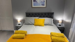 A bedroom with a grey upholstered headboard, a bed with white and yellow bedding, two bedside tables with lamps, and a framed picture on the wall. - New Day Properties-Seaside House-next to beach (South Shields)