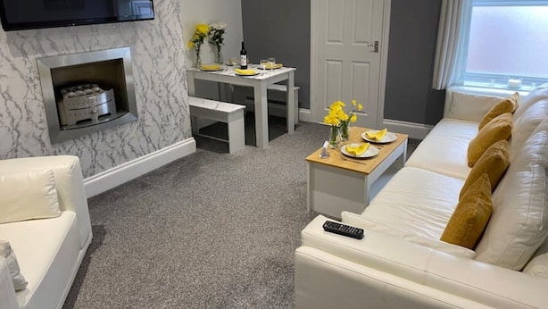 A modern living room with a white sofa, a flat-screen TV mounted on the wall, a dining table with yellow flowers, and a white door. - New Day Properties-Seaside House-next to beach (South Shields)