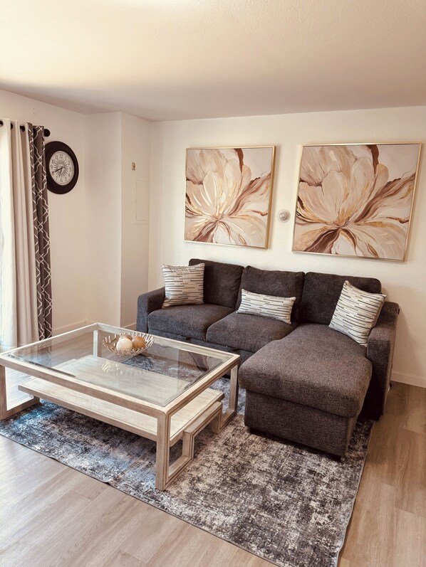 A modern living room with a glass coffee table, two grey sofas, and abstract floral wall art. - North Star Place (Fairbanks)