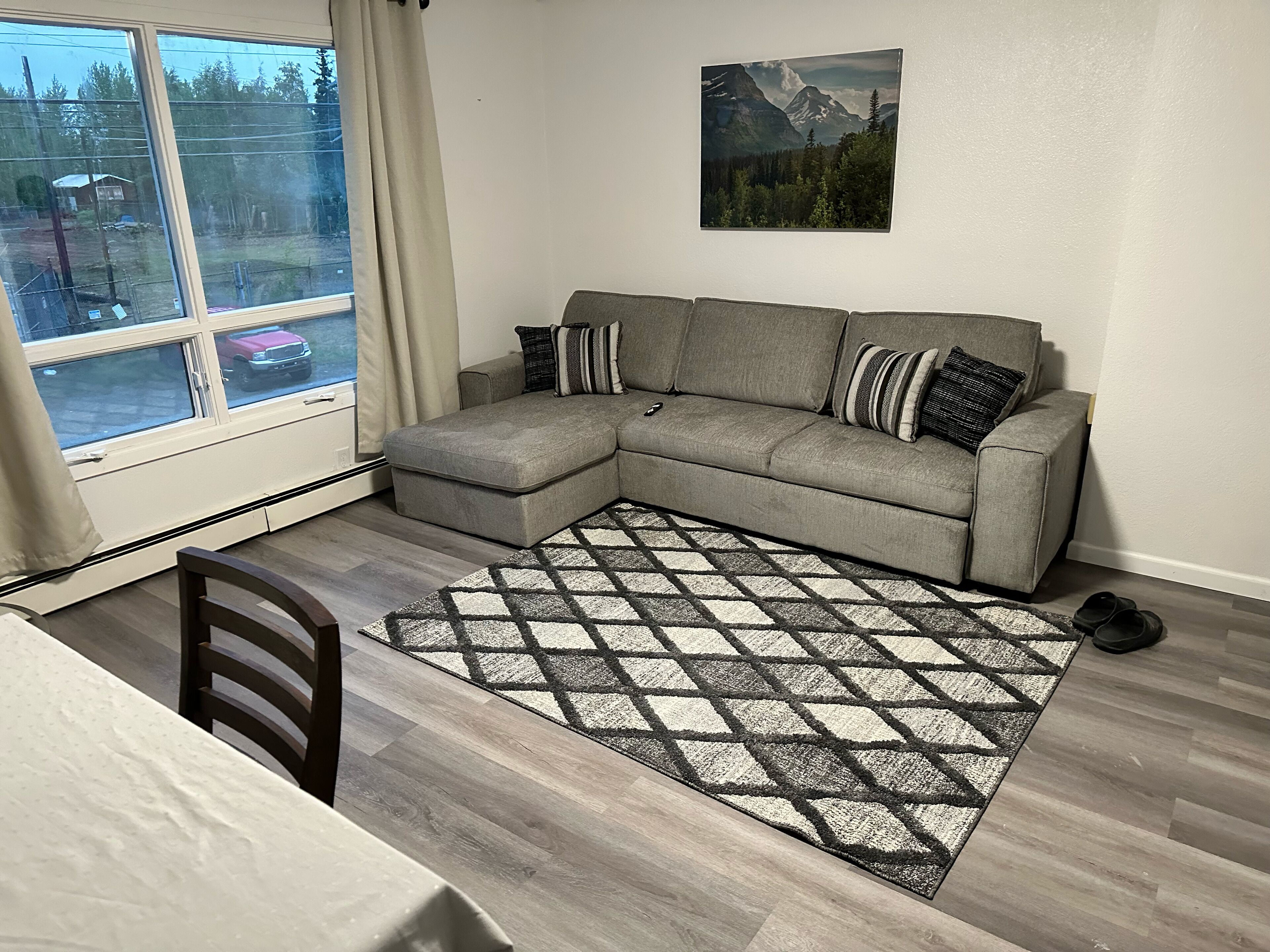A living room with a grey sectional sofa, a patterned rug, a dining table, and a window with curtains.