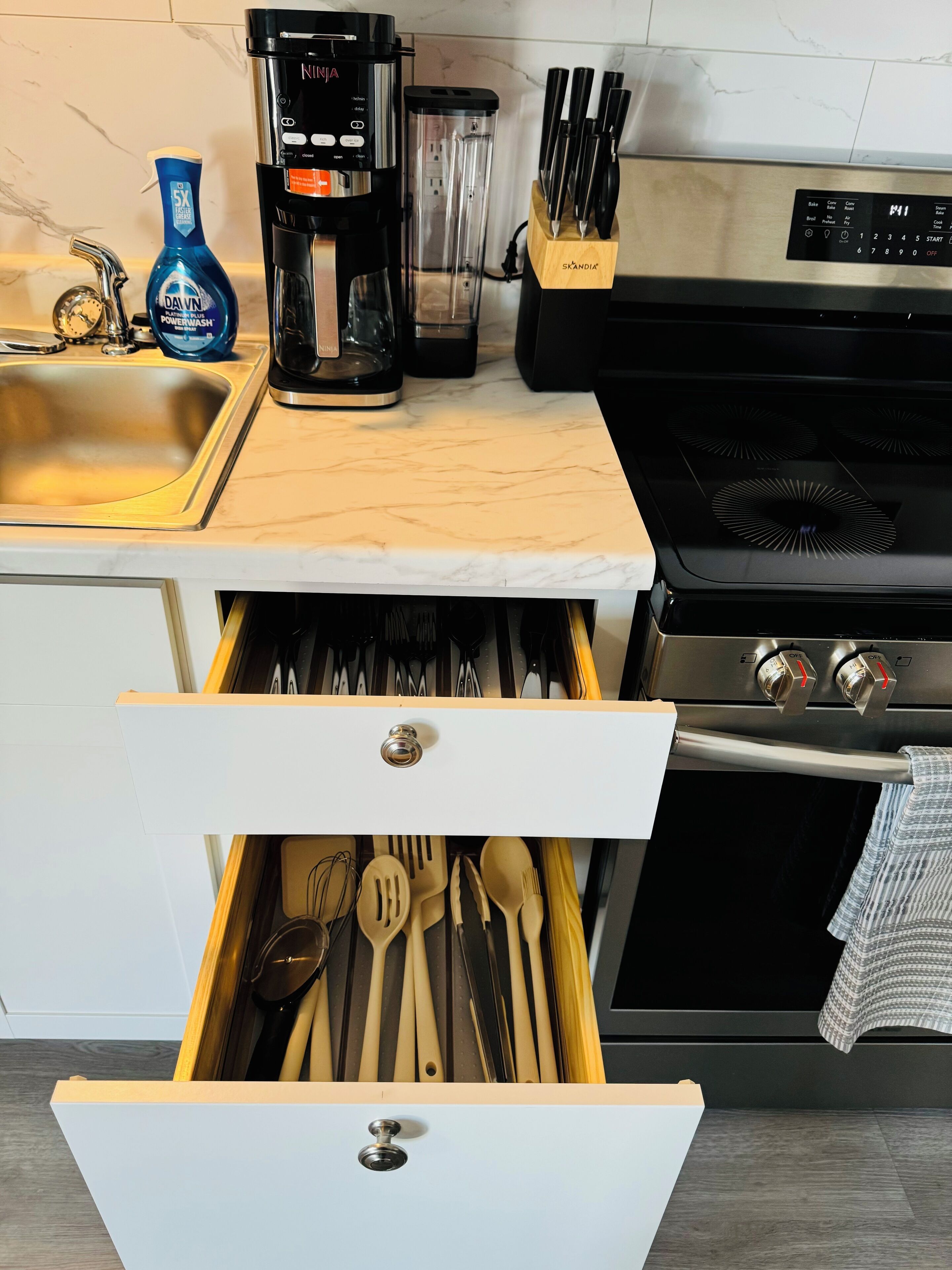 Una cocina con encimera de mármol, cafetera, licuadora y un cajón con utensilios de cocina.