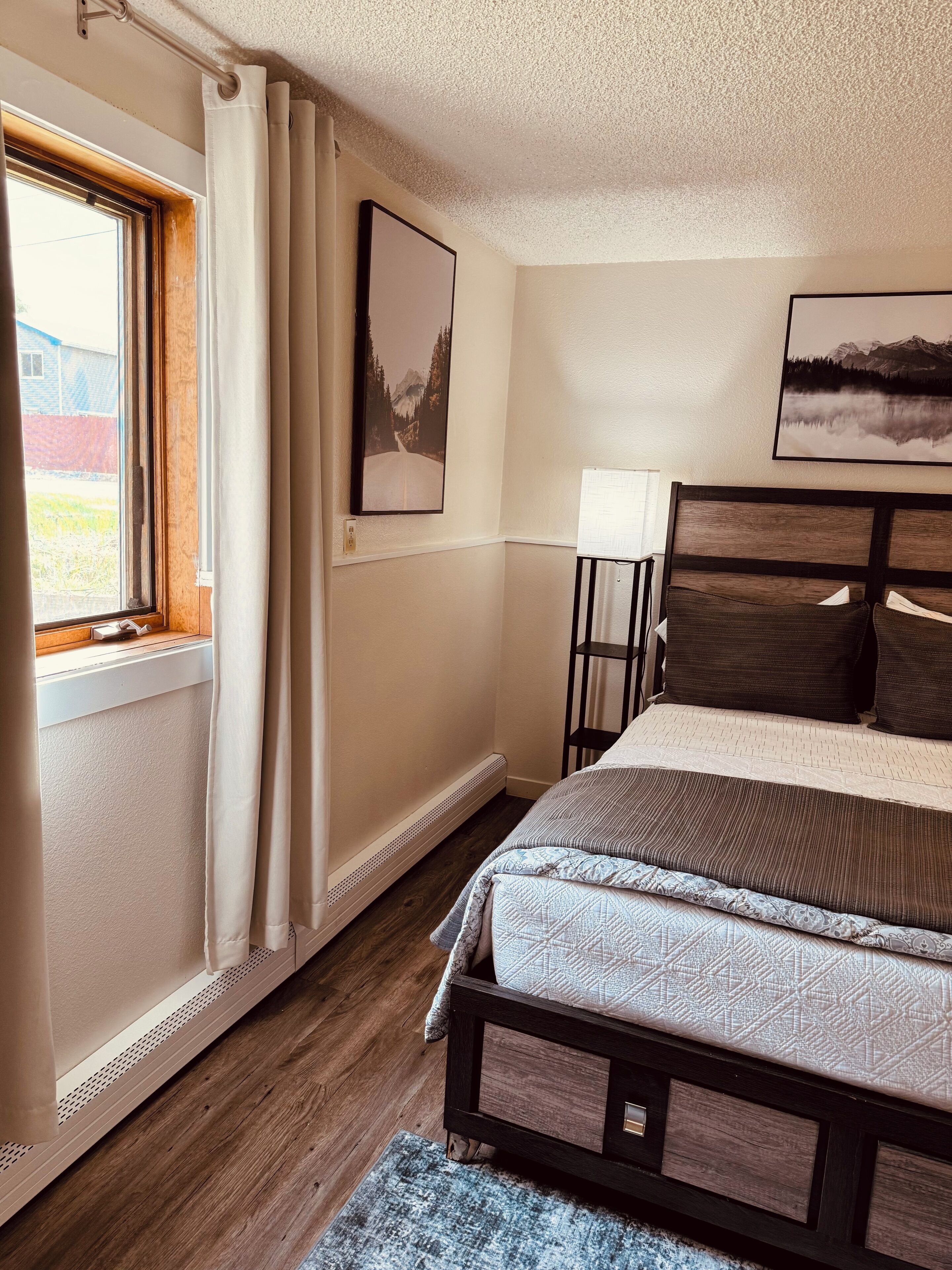 A bedroom with a wooden bed, a window with curtains, and two framed pictures on the wall.
