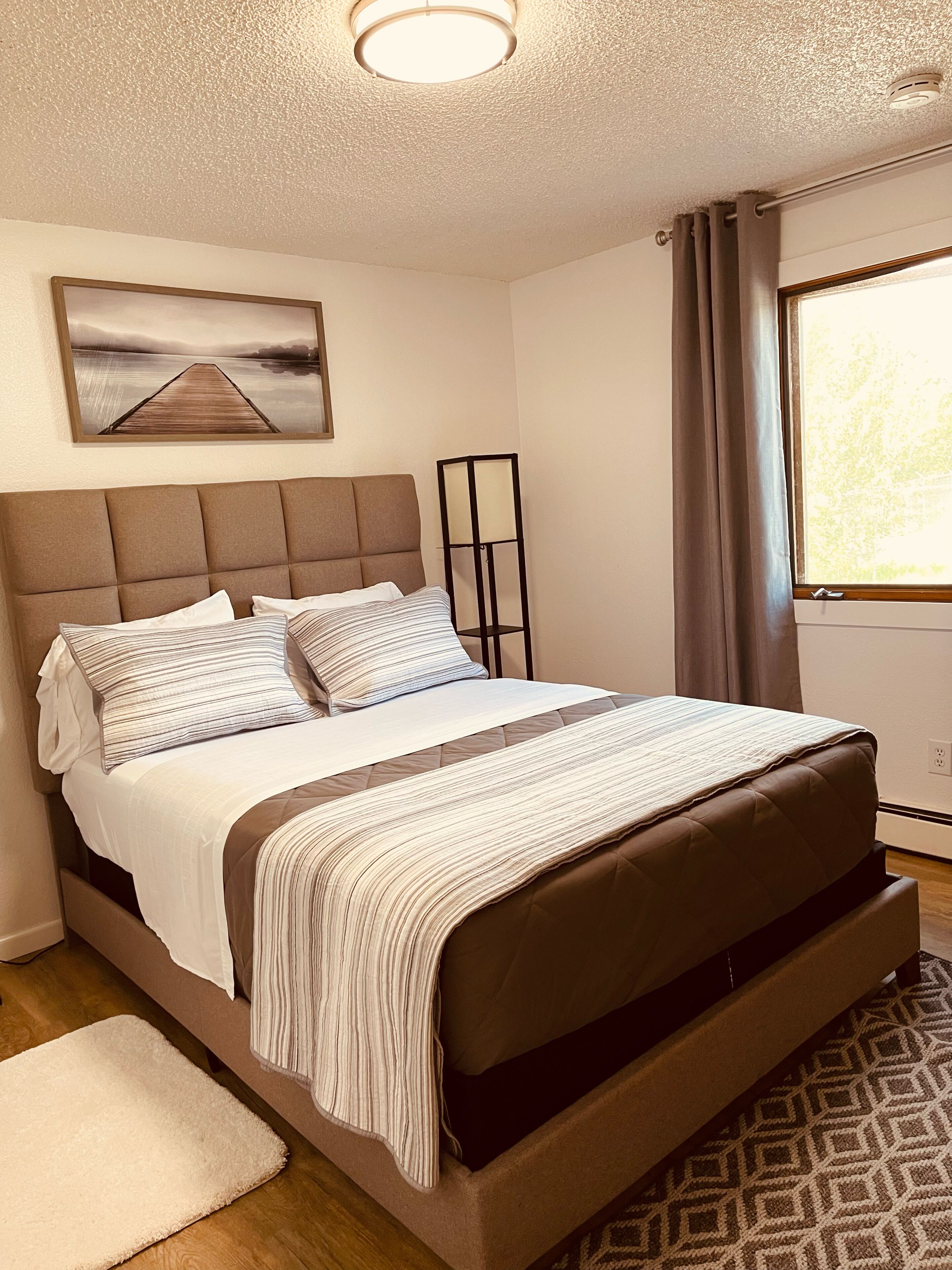 A neatly arranged bedroom with a large bed, a bedside table, a floor lamp, and a window with curtains.