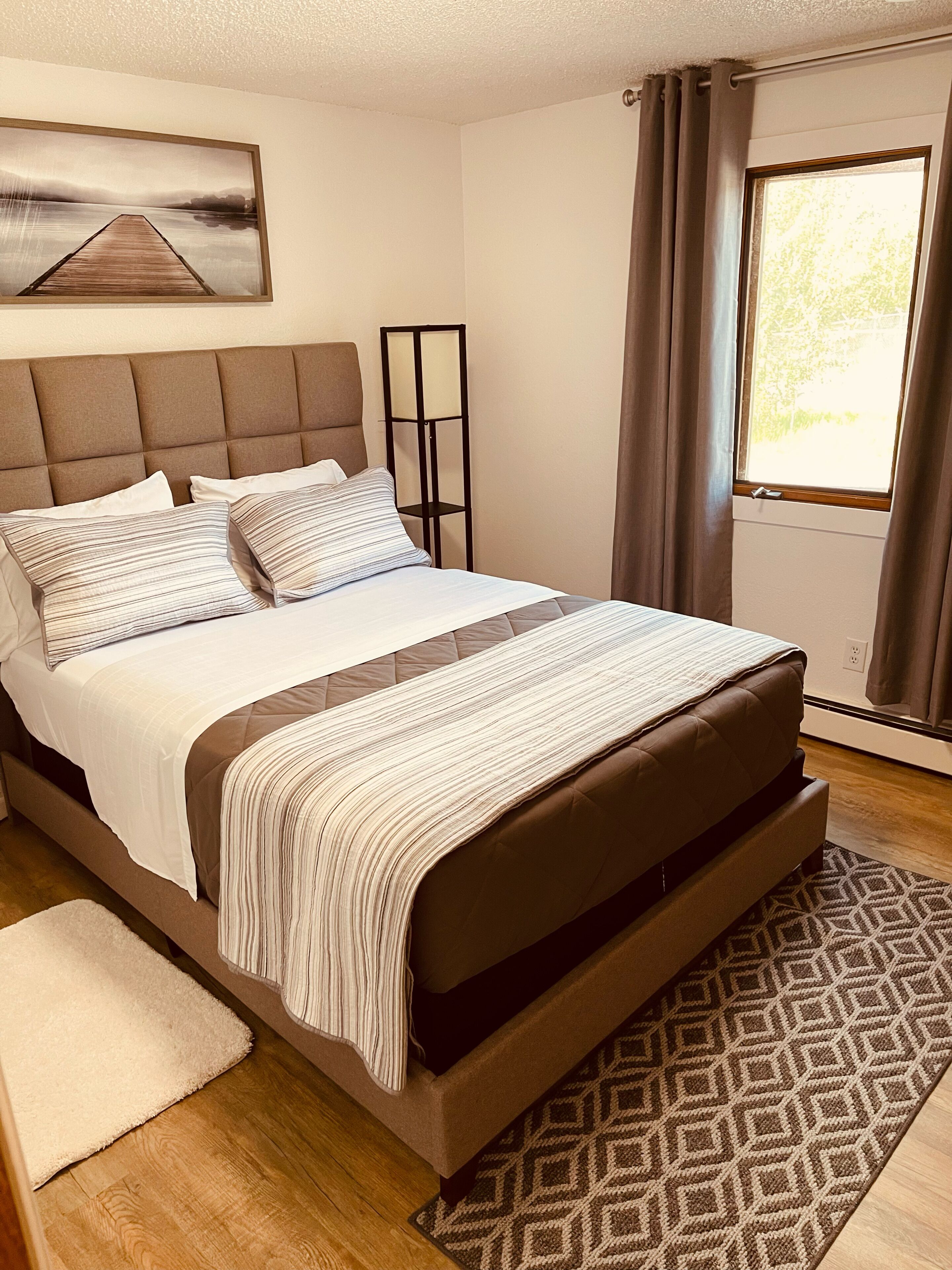 Une chambre à coucher avec un grand lit, une tête de lit, une table de chevet, une lampe sur pied, une fenêtre avec des rideaux et un tableau accroché au mur.