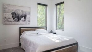 Een slaapkamer met een bed, nachtkastje, raam en een aan de muur hangend schilderij.