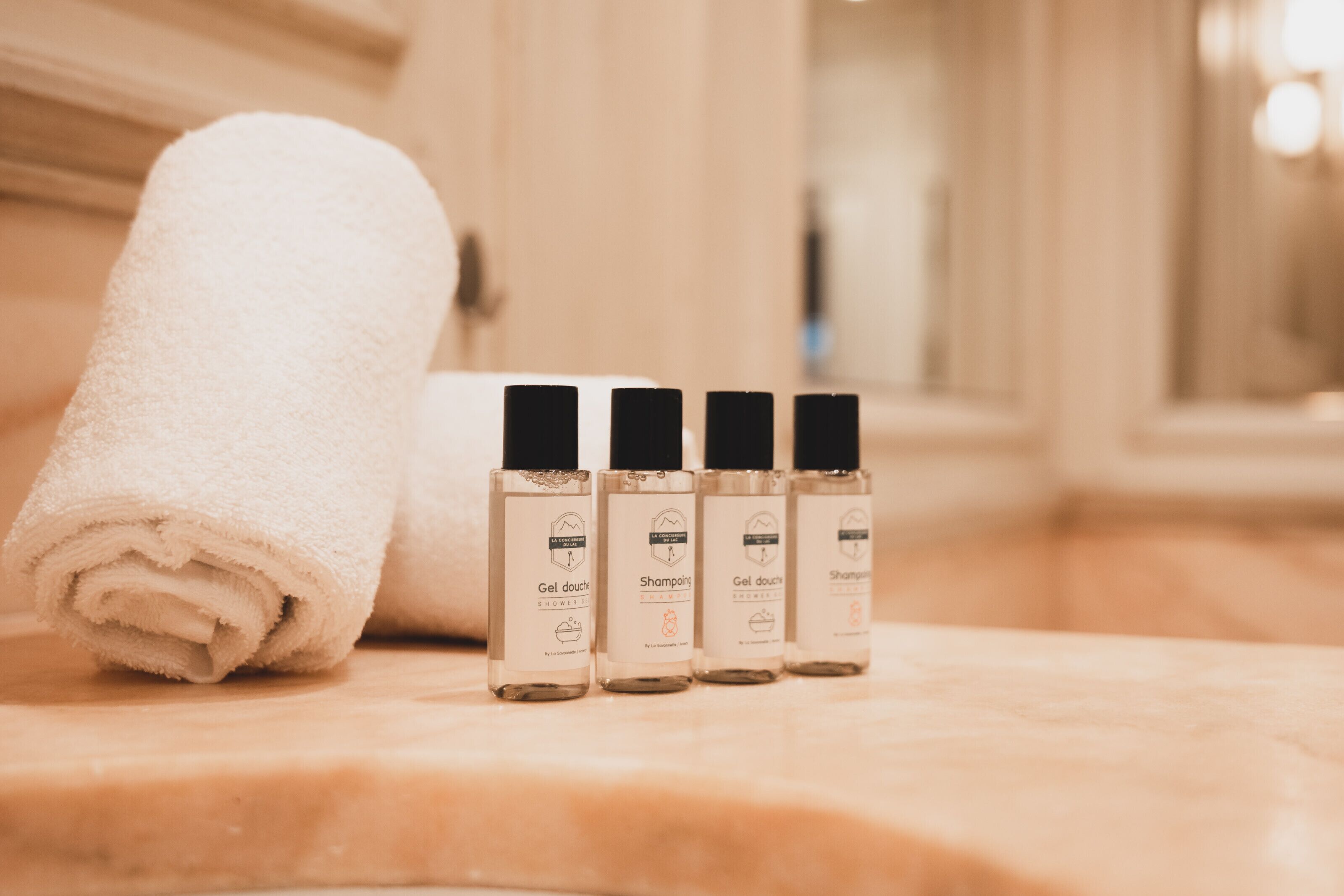 A bathroom with a marble countertop, a rolled white towel, and four bottles of gel douche and shampoo.
