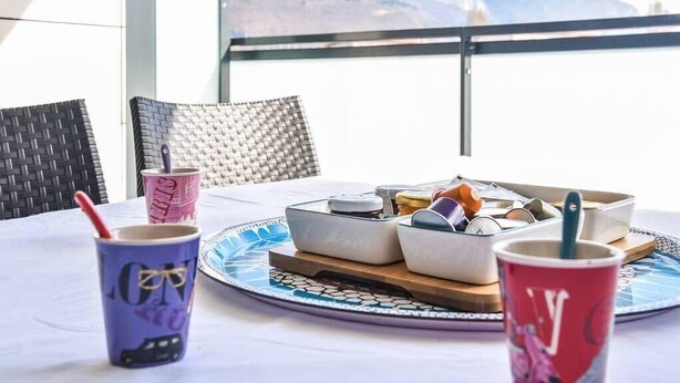 Una mesa con una bandeja de aperitivos, dos tazas con logotipos y vistas a montañas a través de una ventana.