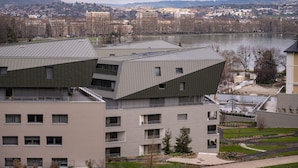 A modern building with a unique angular roof design, multiple balconies, and a view of a lake and distant cityscape. - Le 305 - T3 with garage and beautiful terrace (Annecy)