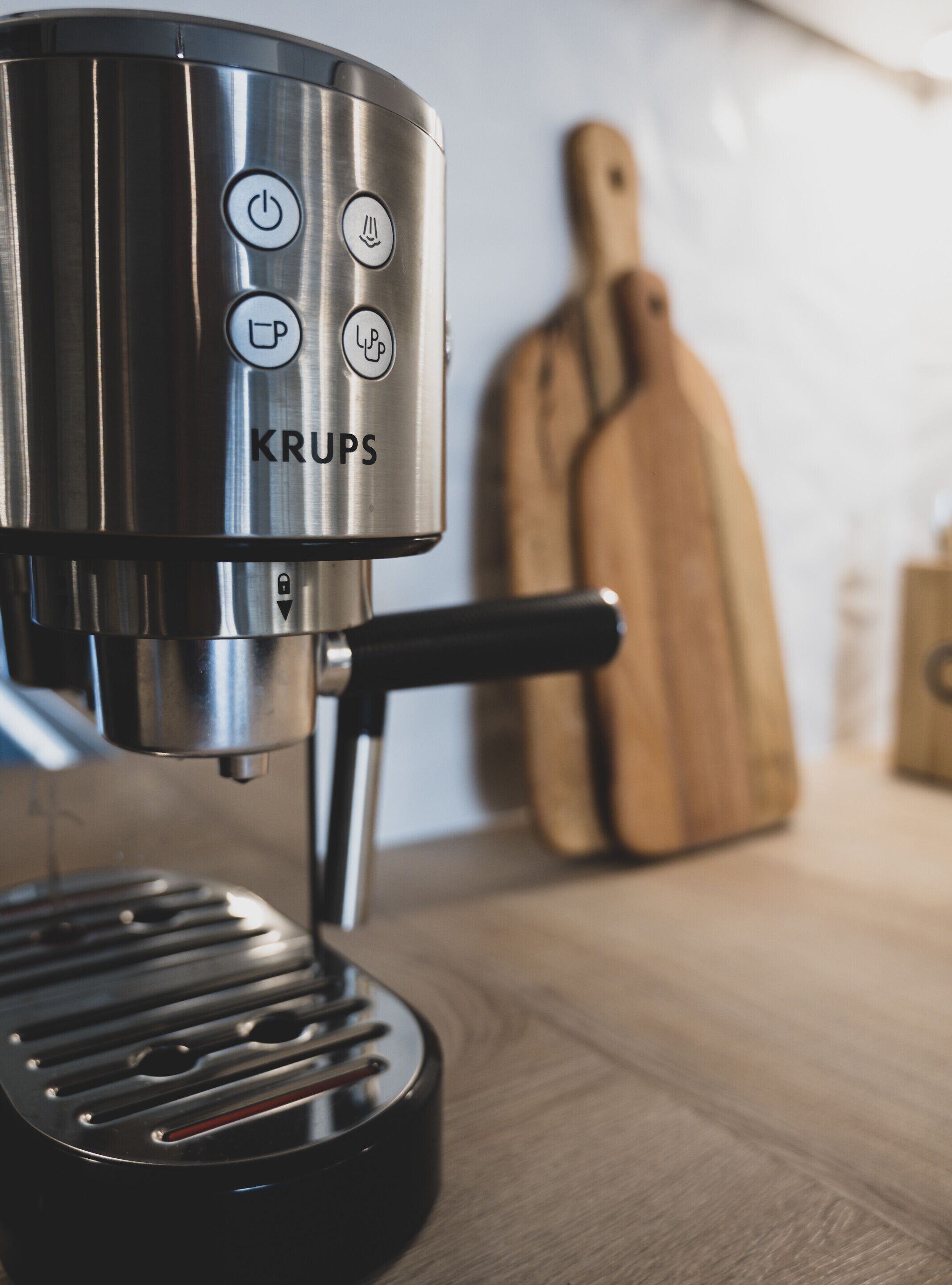 A Krups coffee machine with a black drip tray and a wooden cutting board in the background.