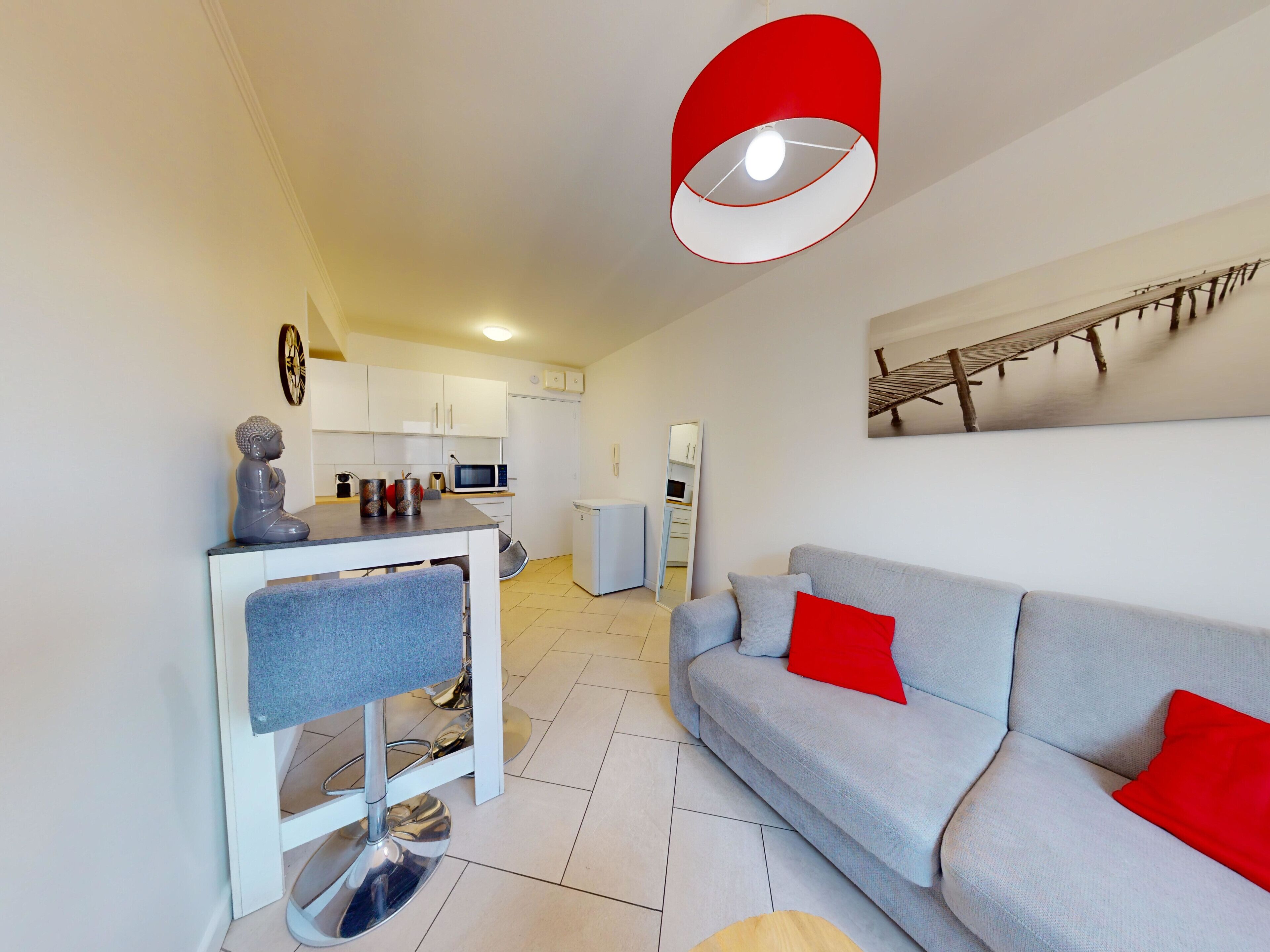Een moderne woonkamer met een grijze bank en rode kussens, een eethoek met een witte tafel en stoelen, en een keuken met witte kasten en een rode hanglamp.