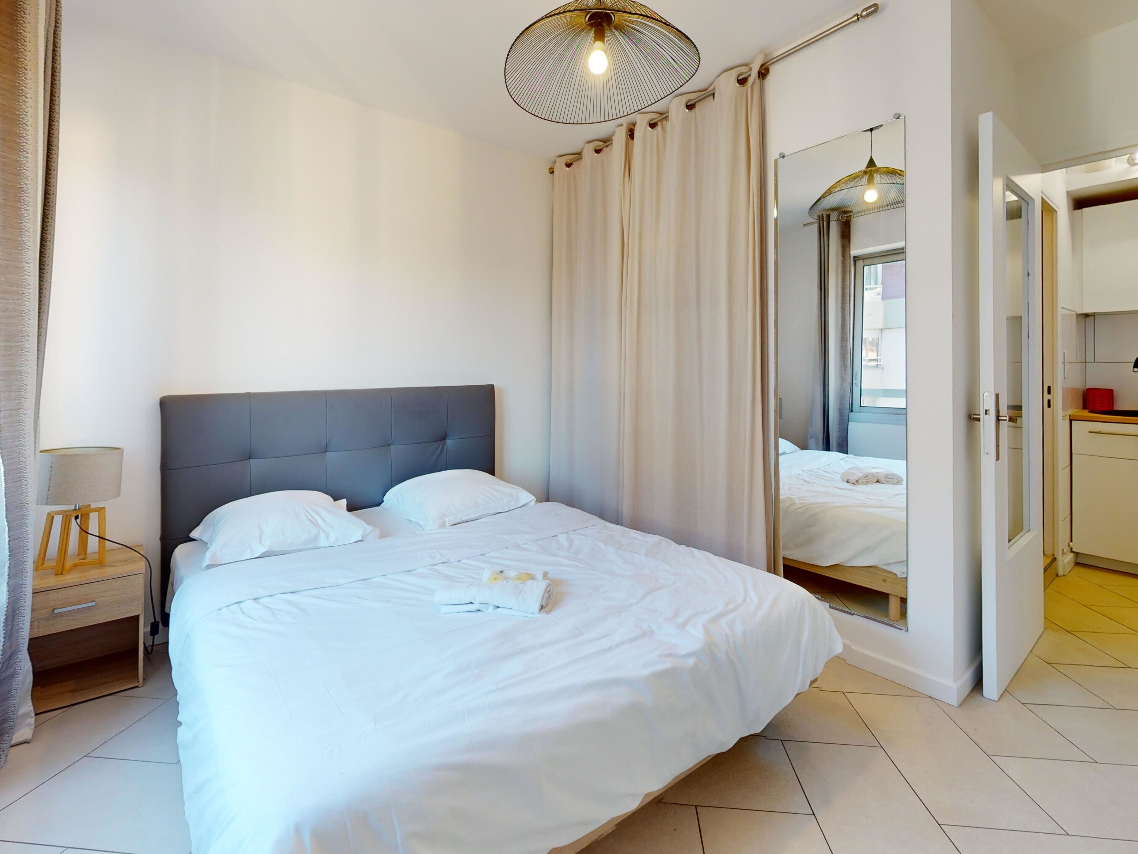 Una cama bien hecha con ropa blanca, un cabecero tapizado en gris y una lámpara de noche.