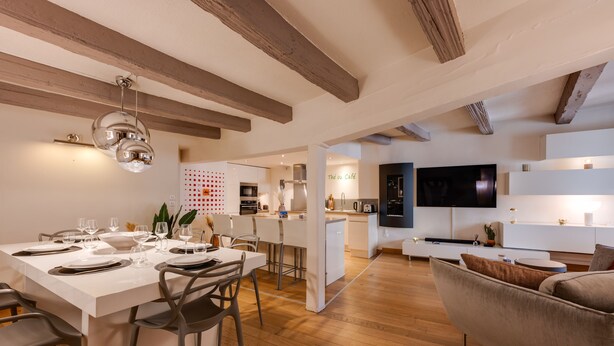 Una cocina moderna con armarios blancos, un comedor con mesa blanca dispuesta para cuatro personas, y una sala de estar con sofá y televisor de pantalla plana.