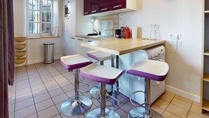 A kitchen with a wooden countertop, a sink, and a window. There are three bar stools with purple seats and chrome bases. A trash can is visible, and a clock is mounted on the wall.