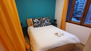 Une chambre à coucher avec un lit, une table de chevet, une fenêtre donnant sur des immeubles et un rideau.