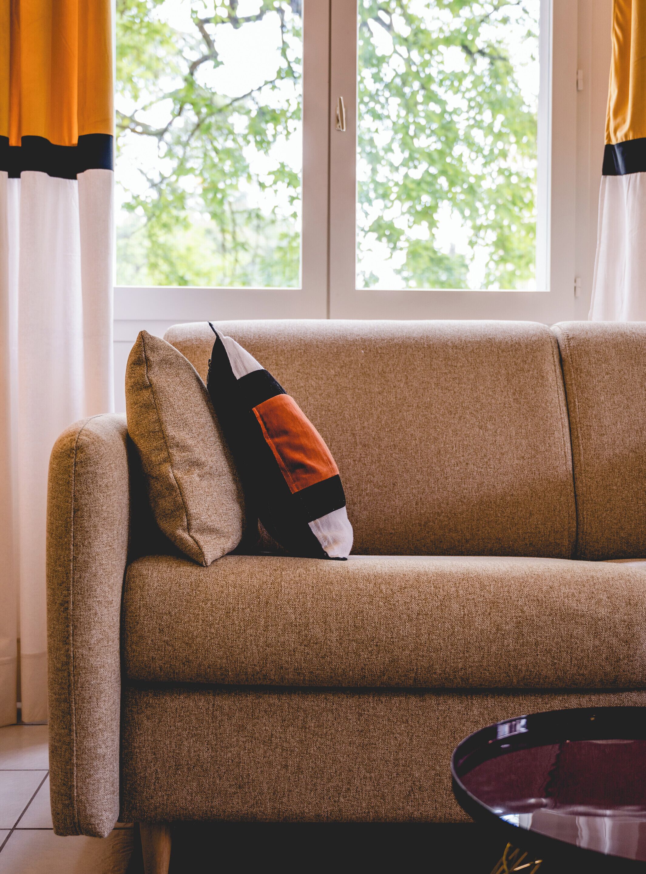 A beige sofa with a single decorative pillow, positioned in front of a window with a view of trees.