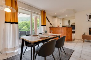 A dining area with a table set for four, a kitchen in the background, and a balcony with curtains.