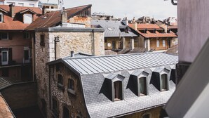 Une vue depuis une fenêtre donnant sur un paysage urbain composé de différents bâtiments, notamment un bâtiment en pierre avec un toit en pente et un bâtiment moderne avec un toit plat.