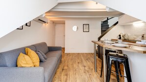 Un espace de vie compact comprenant un canapé gris, un plancher en bois, un coin cuisine avec un tabouret de bar et un escalier.