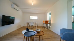 Uma sala de estar moderna com televisão de tela plana, um espaço de refeições com mesa posta para quatro pessoas e uma cozinha visível através de uma porta aberta.