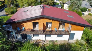 Uma casa moderna de dois andares com telhado vermelho, fachada de madeira e varanda com cadeiras azuis.