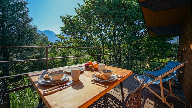 Um deck de madeira com uma mesa posta para o café da manhã, incluindo pratos, xícaras e frutas. Uma cadeira é visível no lado direito.
