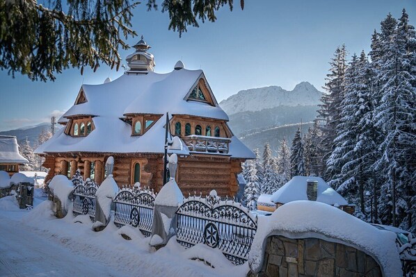 A traditional wooden building with a snow-covered roof, surrounded by snow-laden trees and mountains in the background. - Rich Witch Villa&SPA (Koscielisko)