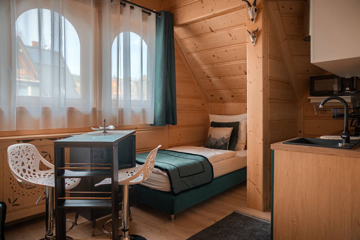Une cabane en bois chaleureuse, avec un lit, une petite table et des chaises, un coin cuisine et de grandes fenêtres avec des rideaux.