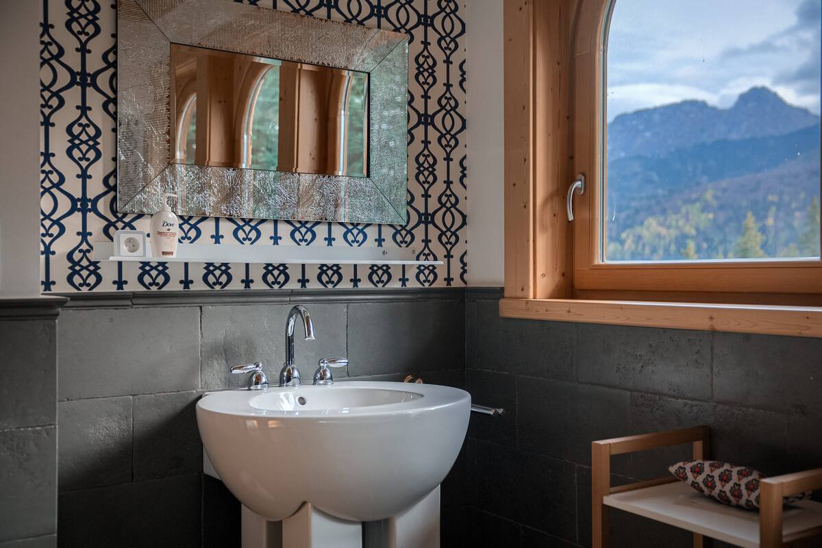 Une salle de bain avec un lavabo blanc, un miroir doté d’un cadre décoratif et une fenêtre donnant sur les montagnes.