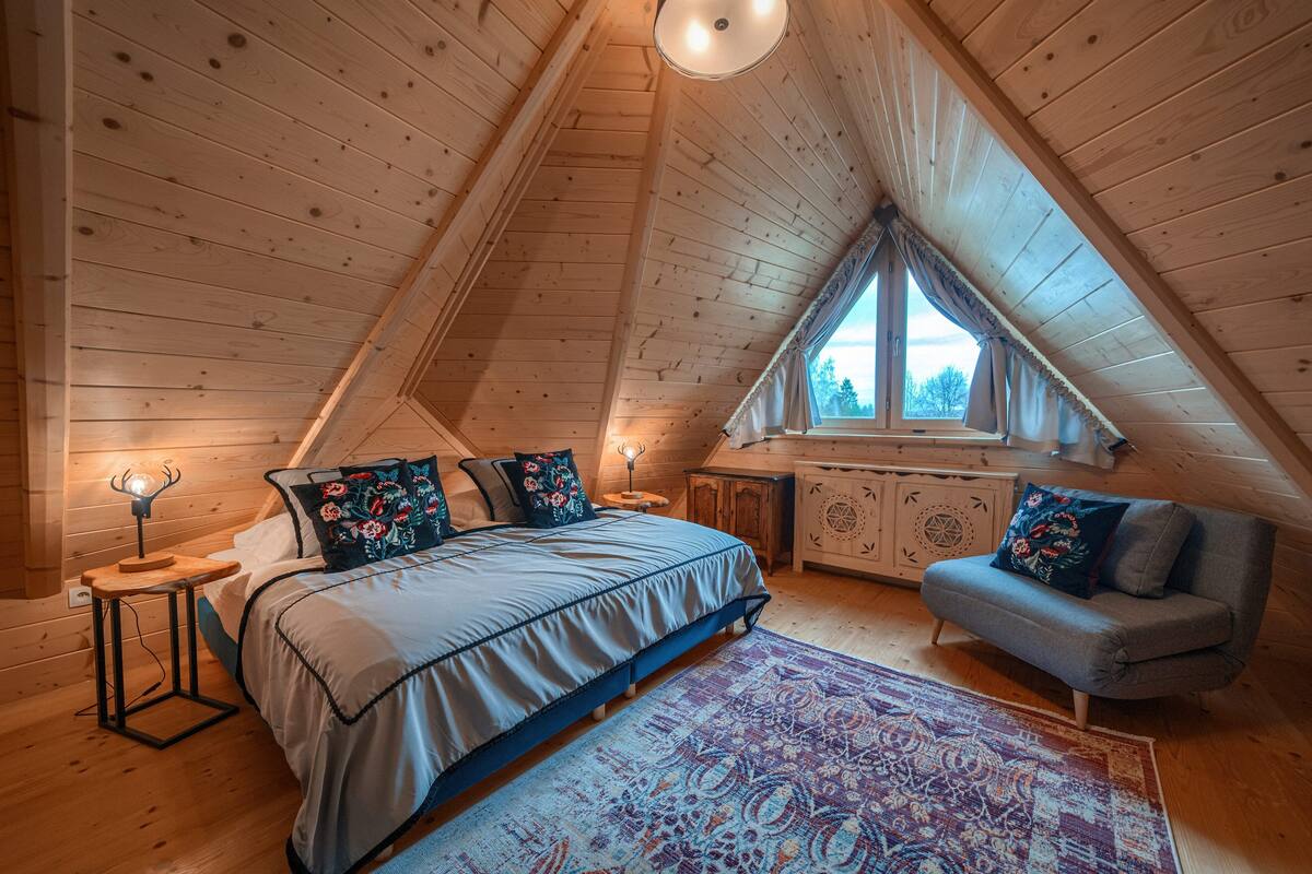 Une chambre confortable située sous les toits, avec un plafond en pente, un lit agrémenté d’oreillers à motifs floraux, une petite table avec une lampe, un banc et une fenêtre avec des rideaux.