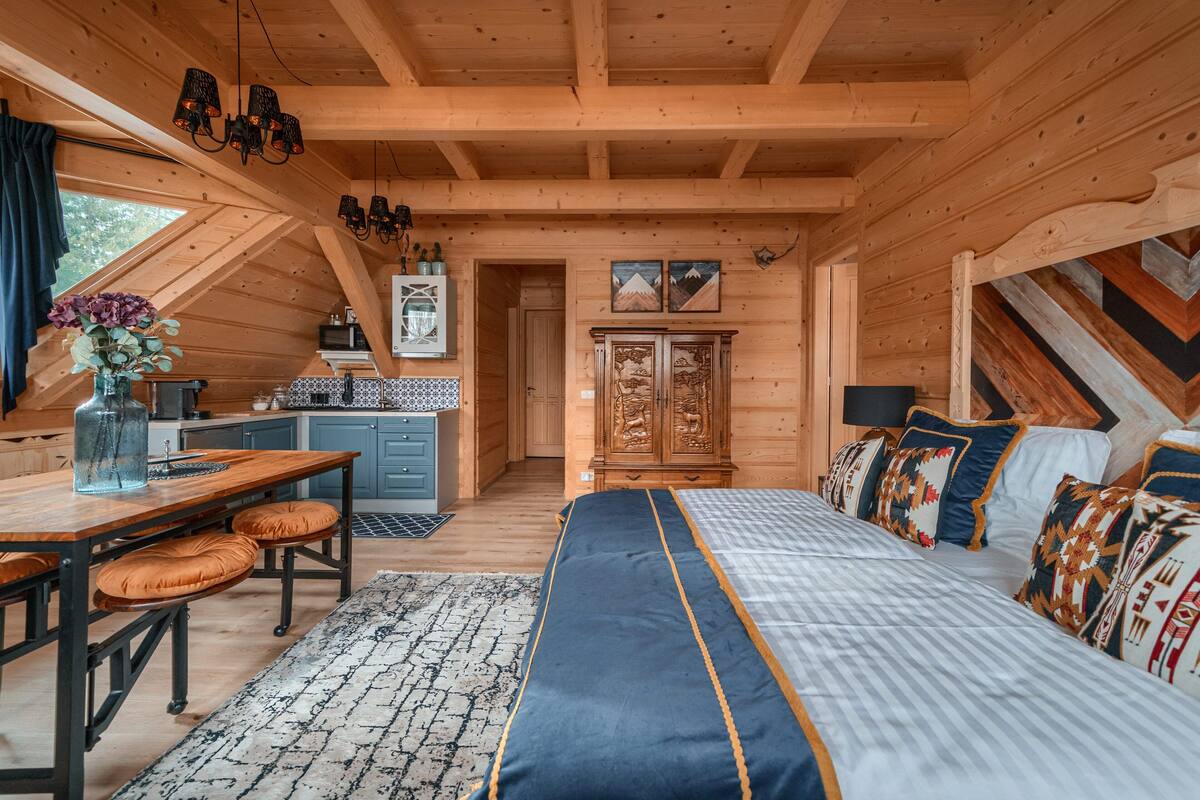 L’intérieur d’un chalet confortable, avec des murs en rondins de bois, une table à manger en bois, un lit agrémenté d’oreillers à motifs et un coin cuisine équipé d’une cuisinière et de placards.