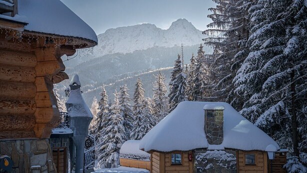 Una cabaña de troncos con techo nevado, rodeada de árboles cubiertos de nieve y montañas al fondo.