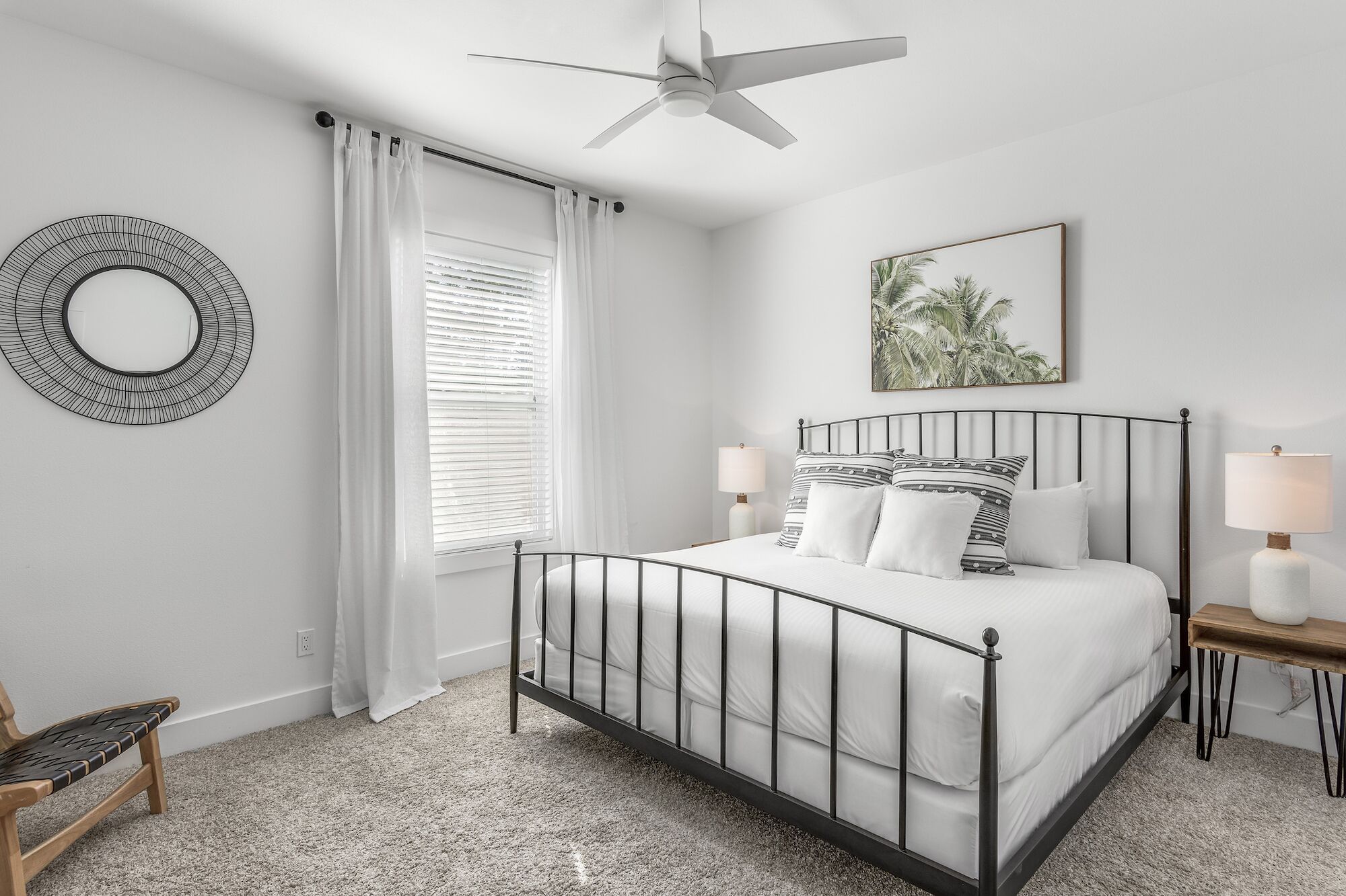 A bedroom with a bed, a ceiling fan, a window with blinds, a bedside table with a lamp, and a wall-mounted mirror.