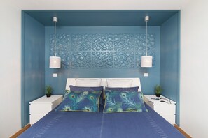 A bedroom with a blue wall, a decorative headboard, two bedside tables with lamps, and a bed with blue bedding and decorative pillows.