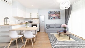 Et moderne køkken med hvide skabe, et spiseområde med et træbord og hvide stole, en grå sofa og et bybillede som kunstværk på væggen.