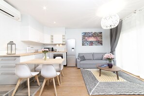 Una cucina moderna con mobili bianchi, un'area pranzo con tavolo e sedie di legno, un divano grigio e un quadro raffigurante un panorama urbano.