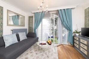 Ein Wohnzimmer mit grauem Sofa, einem weißen Couchtisch, einem Fernseher auf einer Kommode und Blick durch eine Glastür auf einen Balkon mit blauen Vorhängen.