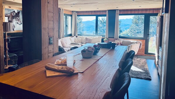 A spacious living area with a long wooden table, comfortable seating, and large windows offering a view of the outdoors.
