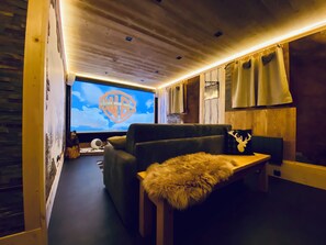 Una habitación acogedora con un televisor de pantalla plana grande mostrando el logotipo de Warner Bros. Pictures, un sofá mullido, una manta de piel sintética y una almohada con forma de cabeza de ciervo.