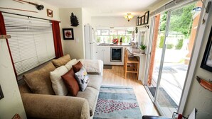 A cozy living room with a sofa, a small kitchen area, and a sliding glass door leading to a garden. - Your next outdoor oasis (Brinnon)