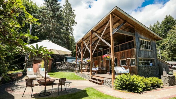 A rustic wooden cabin with a covered patio, outdoor seating, and a stone wall. - Your next outdoor oasis (Brinnon)