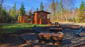 Una baita rustica con facciata in legno, immersa in una zona boschiva. Si notano due baite, un tavolo da picnic e due poltrone Adirondack.