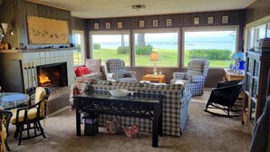 A cozy living room with a fireplace, plaid furniture, and large windows overlooking a scenic view. - 4 guest · Eddie Bauer`s former Beachfront Cottage (Sekiu)