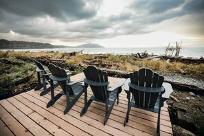 A wooden deck with four Adirondack chairs facing a body of water, surrounded by natural scenery and a cloudy sky. - The Beach Place | Beachfront, Pet-Friendly, Views (Port Angeles)
