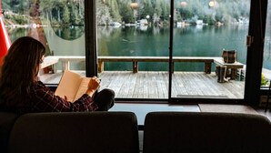 Una persona leyendo un libro junto a una ventana con vistas a un lago y un bosque.