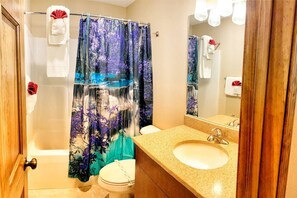 Une salle de bain avec un rideau de douche représentant une scène de forêt, des toilettes, un lavabo avec un miroir et des serviettes accrochées au mur.