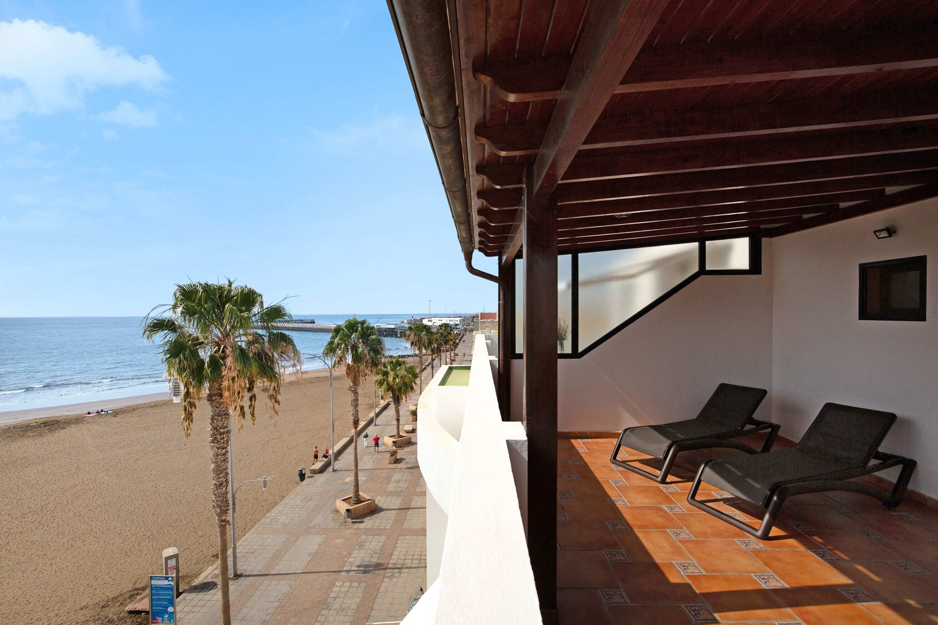 Un balcón con dos sillones de descanso, vista a la playa, palmeras y el océano.