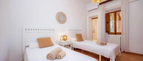A hotel room with two beds, a bedside table with a lamp, a mirror, and a window with curtains.