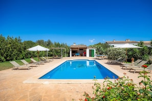 Une piscine entourée de verdure, agrémentée de chaises longues et de parasols, et bordée d’un bâtiment dont le toit est recouvert de tuiles.