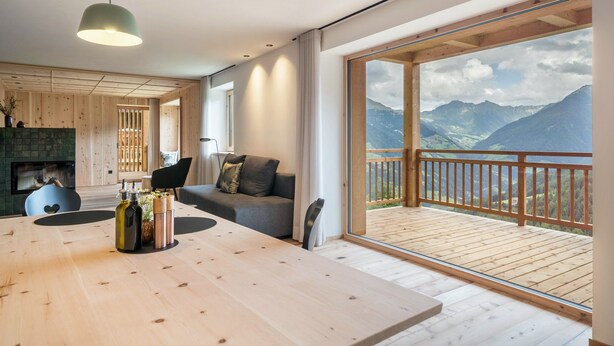 A modern interior with a wooden dining table, a sofa, and a balcony with a mountain view. - Apartment 'Mucher Veit' with Mountain View, Shared Garden and Wi-Fi (Moso in Passiria)