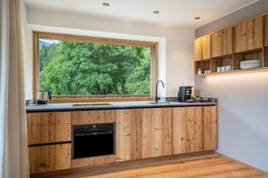 A modern kitchen with wooden cabinetry, a built-in oven, a sink, and a window overlooking a forest.